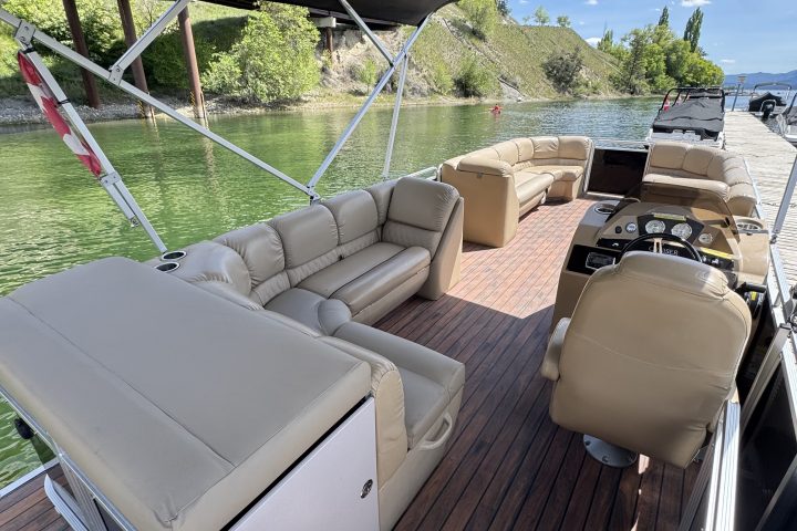 Interior of a pontoon boat with beige seating, under a canopy, docked by a scenic lake.