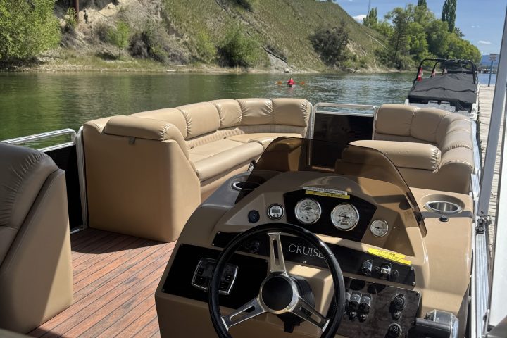 Interior view of a boat with tan seats and a steering wheel, near a riverbank and bridge with trees.