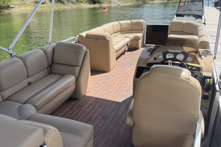 Interior of a boat with beige seating and wooden floor, docked by a scenic green lake.