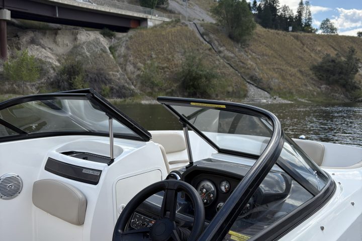 White boat with windshield, steering wheel, and wooden deck near a bridge and hillside.