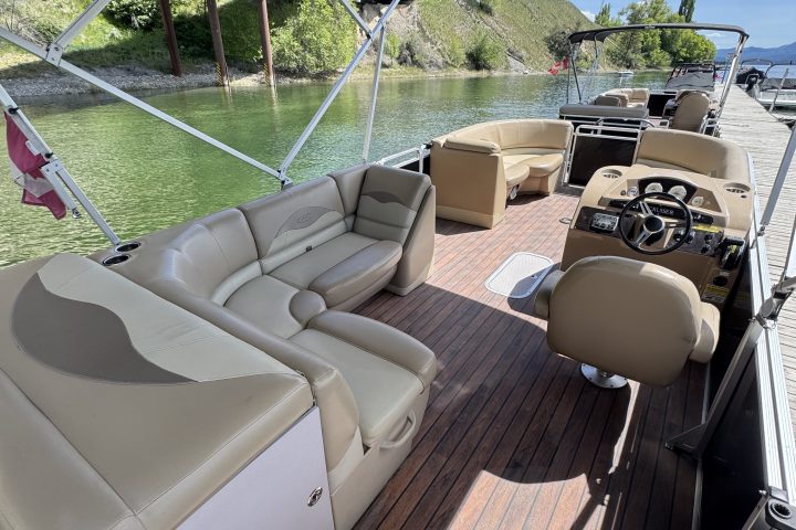View of a pontoon boat's interior with tan seating and a steering console near a lake.